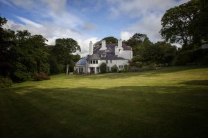 A picture of a large white house surrounded by trees and a lawn. The house is Tŷ Newydd Writing Centre.