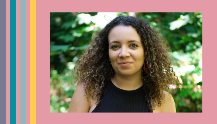 a woman with black curly hair wearing a black top stands in front of some trees