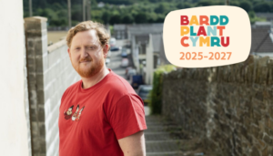 A person in a red t - shirt standing in front of a wall. Text on the picture reads Bardd Plant Cymru 2025-2027.